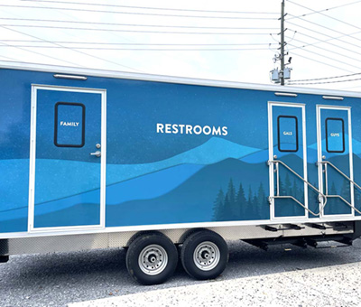 An exterior view of a Lang ADA + 5 Park Series 2 Restroom Trailer with a blue mountain outer wrap applied to the trailer exterior.