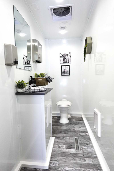 An interior view of a Lang 8 Station Pro Series Restroom trailer with toilet, sink vanity, mirror, and decor.
